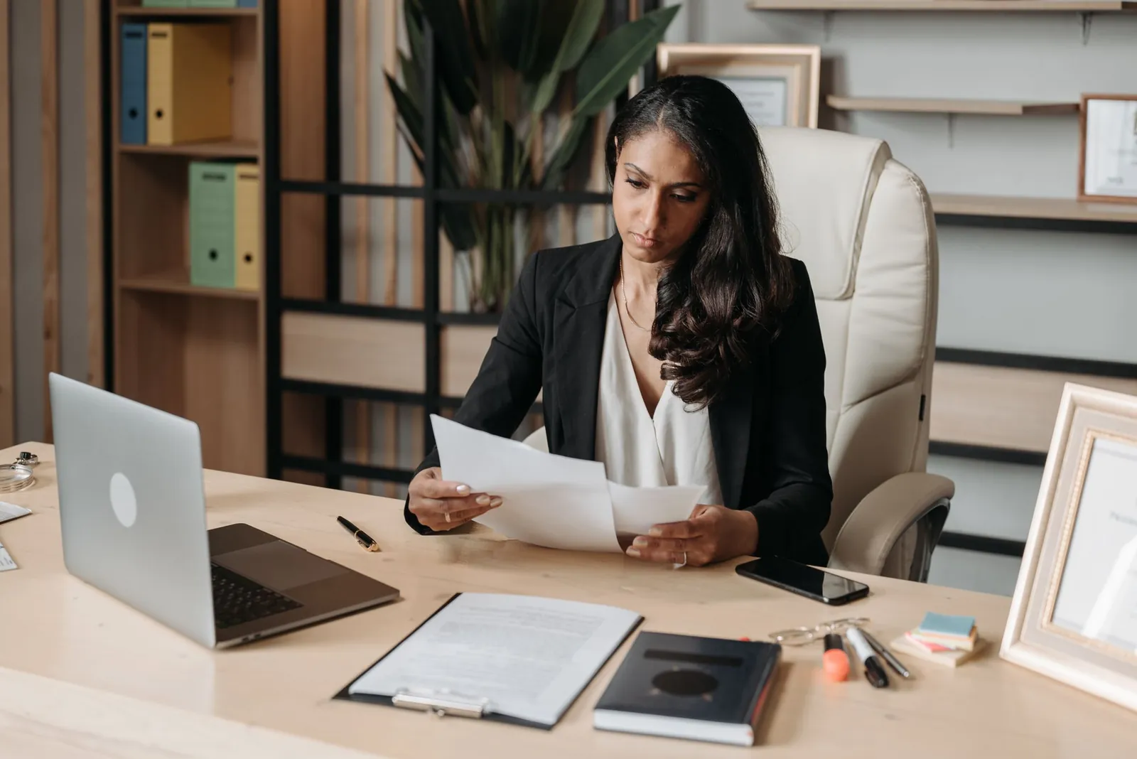 A woman at an office desk working through printed documents, a laptop open beside her.
