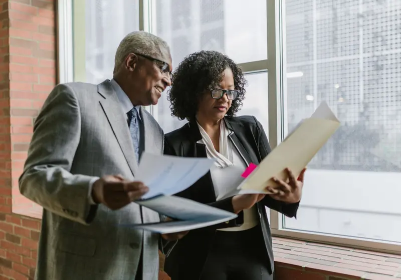 An older man and a woman in business clothes stand by a bright window reading papers together.