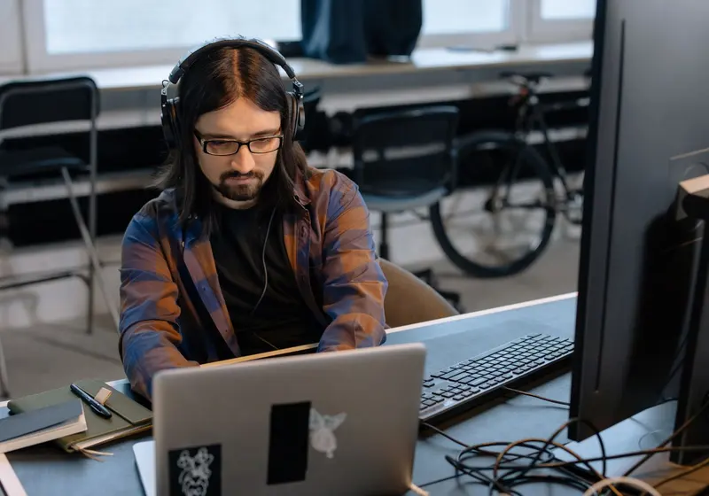 A man with headphones works on a laptop beside a large monitor.