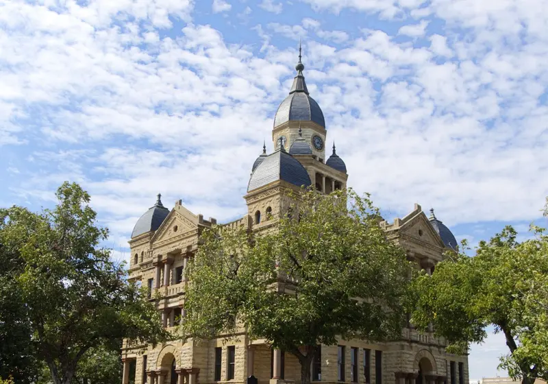 A stone county courthouse with a clock tower stands behind trees on a sunny day.