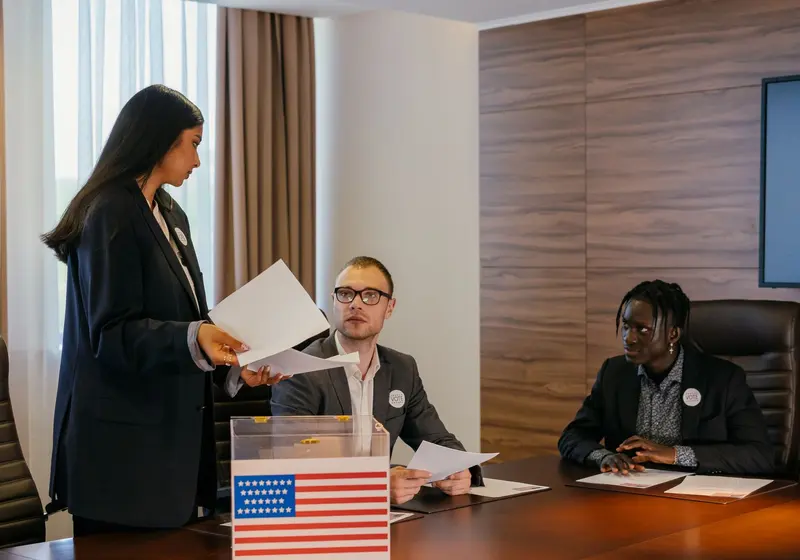 A woman hands papers across a conference table to two seated men beside a ballot box with a flag.