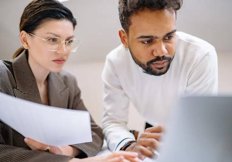 A woman holding a paper and a man leaning over a laptop look at the screen together.