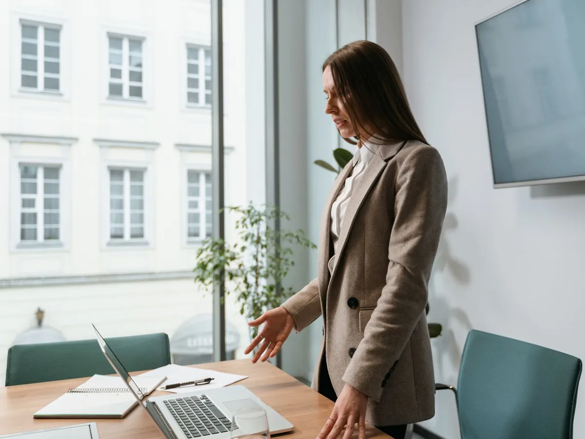 A manager standing at a meeting table with a laptop, by a window onto a stone building.