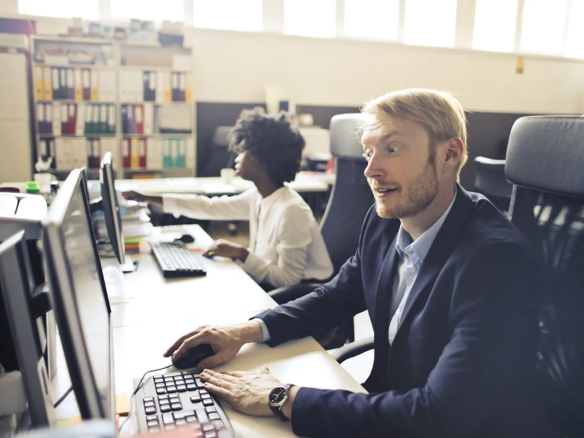A staff member working at two monitors in a bright open office, a colleague at the next desk.