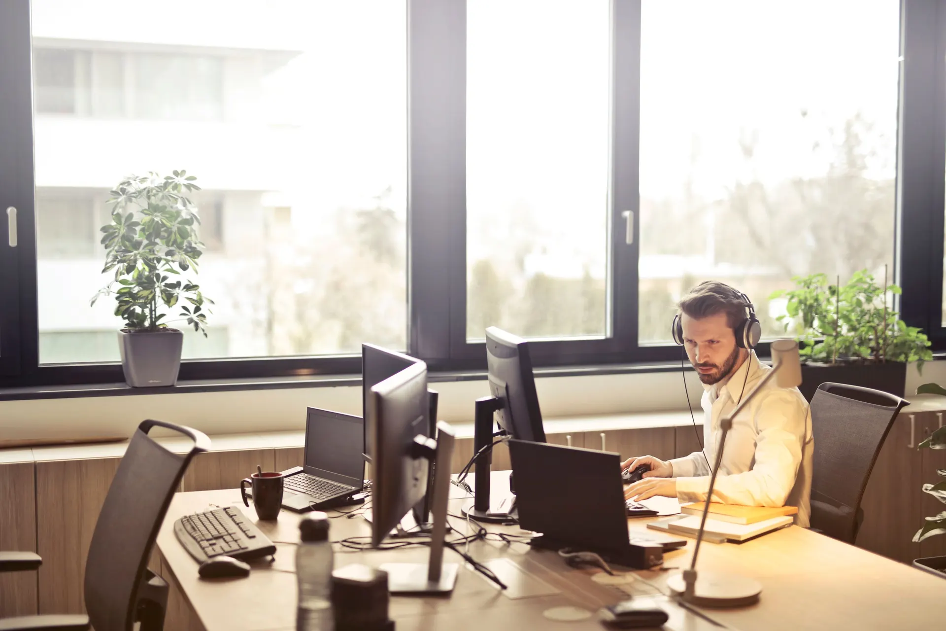 An IT staff member in headphones works at a desk of monitors and laptops in a bright office.
