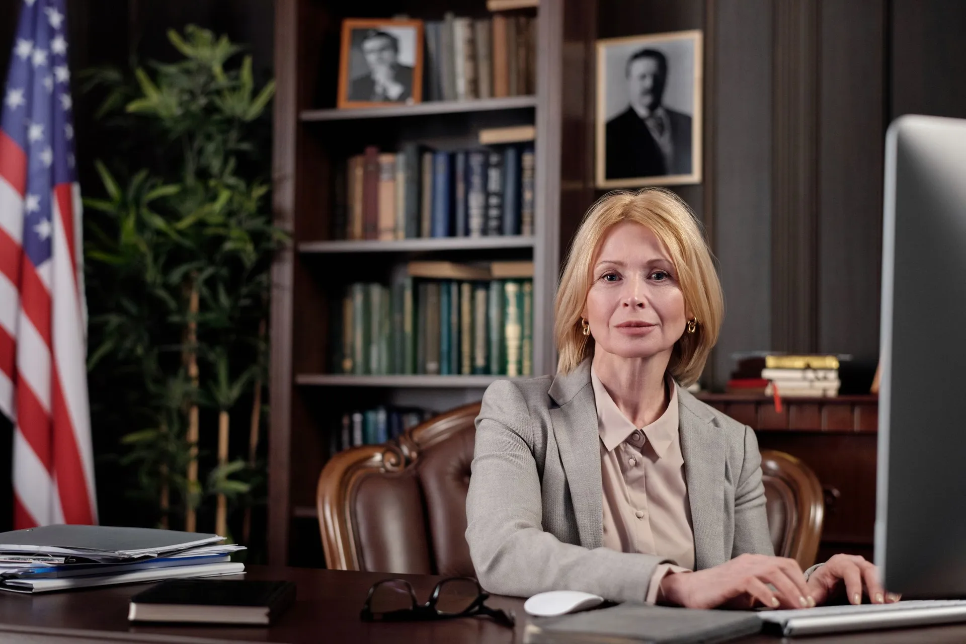 An official works at her desk in a wood paneled office, with a US flag and bookshelves behind her.