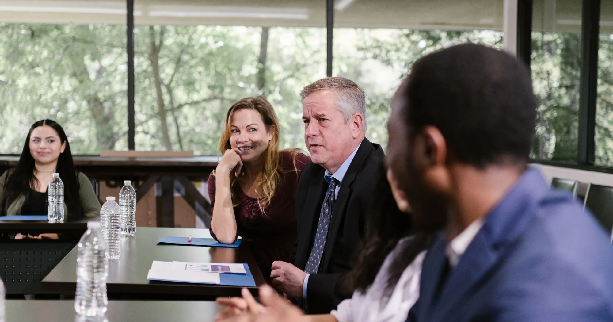 Colleagues around a meeting table by a window, listening as one of them speaks.