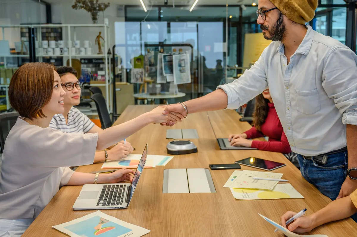 Two people shaking hands across a work table where colleagues sit at laptops.