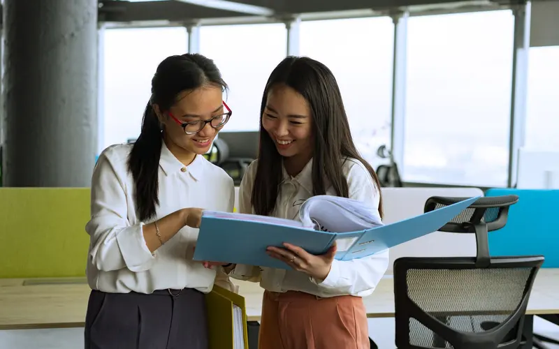 Two women in an office look through a blue binder together.