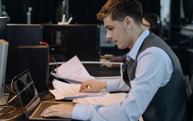 A man at an office desk types on a laptop beside stacks of papers.