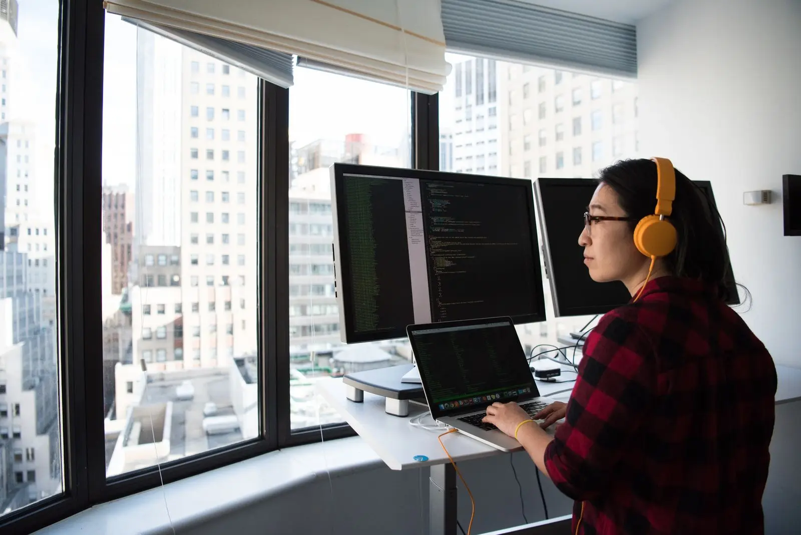 A woman in yellow headphones works at a desk with several monitors beside a bright window.
